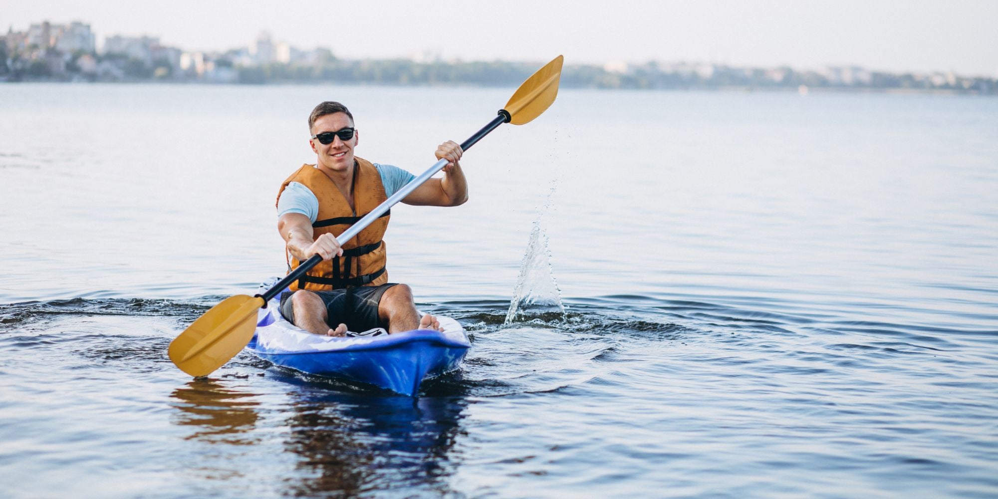 Man paddling a kayak on a calm body of water with a city skyline in the background.
